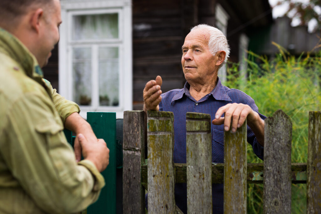 a man in the middle of the image is standing behind a fence that surrounds his house. He is gesturing with his hand facing another man on the left-side of the image.