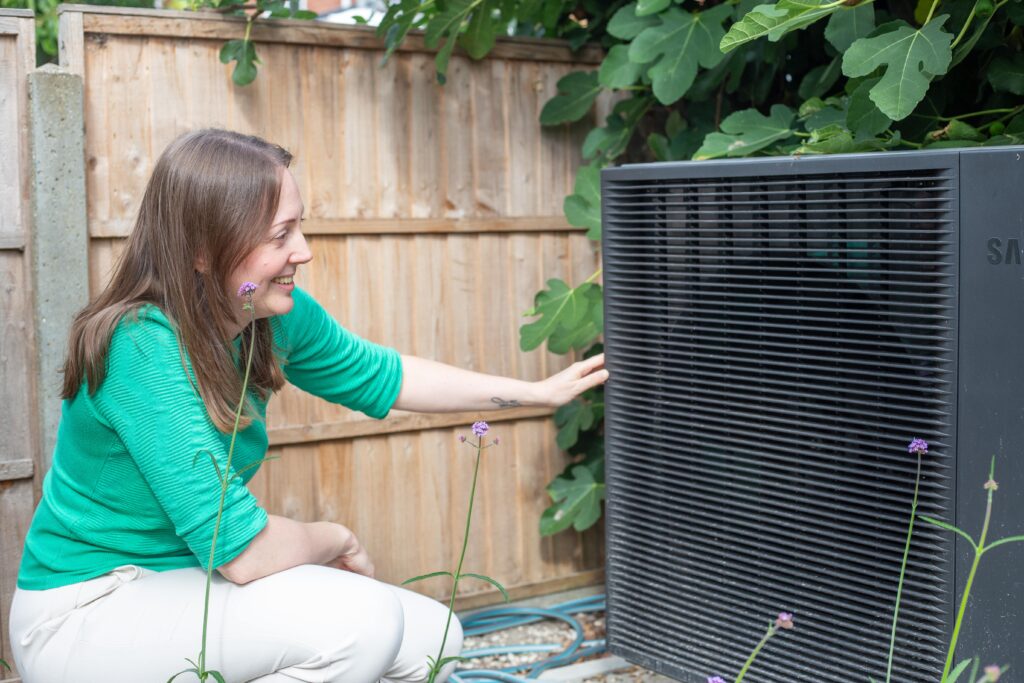 woman squatting down next to a black heat pump in a garden. Her hand is resting on the heat pump as she looks at it.